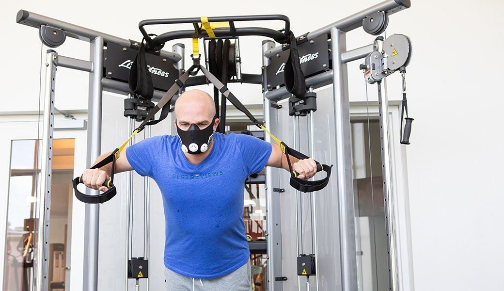A bald person in a blue t-shirt uses exercise equipment while wearing an elevation training mask.